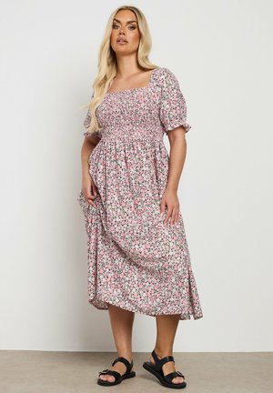 Robe midi à motifs floraux en rose avec des accents verts, décolleté carré, manches courtes bouffantes et corsage froncé. Associée à des sandales noires.