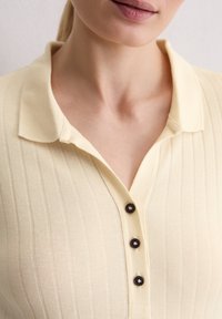 Nahaufnahme einer Person, die ein cremefarbenes, geripptes Poloshirt mit drei schwarzen Knöpfen und einem Kragen trägt, wobei der Hals und die untere Gesichtshälfte zu sehen sind.