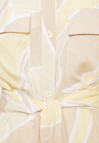 Robe-chemise beige clair et jaune à motifs, avec des boutons sur le devant, deux poches poitrine avec rabats, et une ceinture en tissu nouée à la taille.