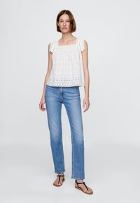 Top bianco senza maniche in cotone con ricamo ad occhiello e bordo smerlato, abbinato a jeans denim azzurri e sandali neri bassi.