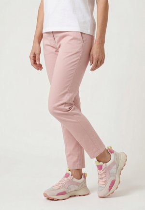 Osoba w jasnoróżowych obcisłych spodniach i białoróżowych tenisówkach stoi z jedną nogą lekko ugiętą na jednolitym tle.