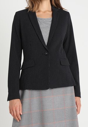 Femme portant un blazer noir à pois boutonné à la taille par-dessus un haut gris et une jupe grise à carreaux avec des lignes rouges.