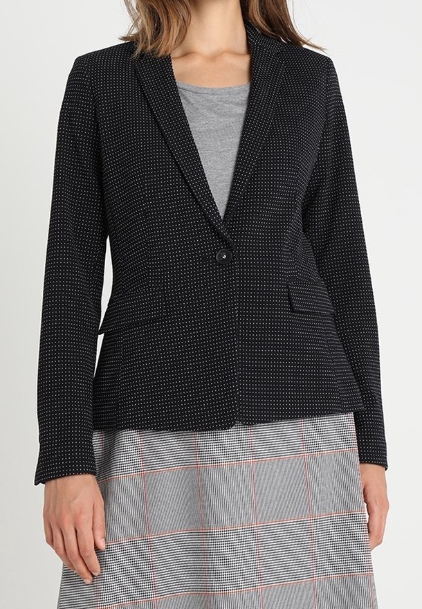 Femme portant un blazer noir à pois boutonné à la taille par-dessus un haut gris et une jupe grise à carreaux avec des lignes rouges.