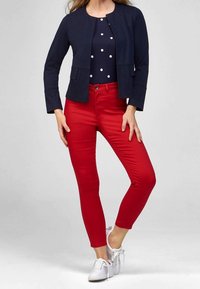 Nainen, jolla on yllään tummansininen takki, tummansininen paita valkoisilla pilkuilla, punaiset slim-fit-housut ja valkoiset lenkkarit, seisoo yksinkertaista vaaleaa taustaa vasten.