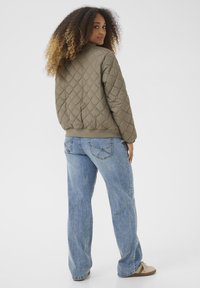 Veste bomber matelassée vert olive avec des poignets et un ourlet côtelés, associée à un jean en denim bleu clair. Modèle de dos, mettant en avant le design arrière.