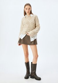 Maglione lavorato a maglia beige con motivi a rombi, indossato sopra una camicia bianca. Gonna in tessuto herringbone marrone e stivali alti marroni con fibbie.