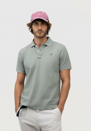 Hombre con una camiseta polo gris claro, pantalones de color claro y una gorra de béisbol rosa desvaída, de pie con las manos en los bolsillos.