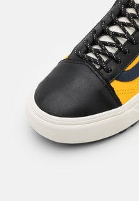 Sapatilha de couro preta com ponta arredondada, detalhes em amarelo e azul-marinho, sola de borracha texturizada e atacadores pretos e brancos.