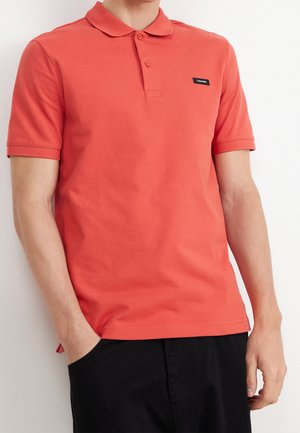 Mężczyzna ubrany w koralową koszulkę polo z krótkim rękawem z małym czarnym logo Calvin Klein na piersi, ręce w kieszeniach czarnych spodni.
