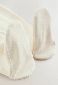 Nahaufnahme von weichen, cremefarbenen Babyhandschuhen, die flach auf einer hellen Oberfläche liegen, und dabei ihre glatte Stoffstruktur und die runde Form hervorheben.