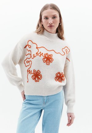 Pull en tricot blanc avec broderie florale orange, doté d'un col côtelé haut et de poignets côtelés, porté avec un jean bleu clair.