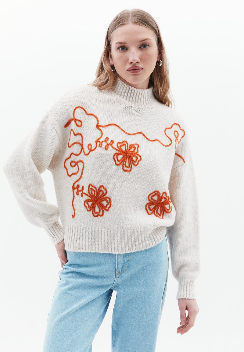 Maglione bianco a maglia con ricami floreali arancioni, caratterizzato da un alto colletto a costine e polsini a coste, indossato con jeans azzurri chiari.