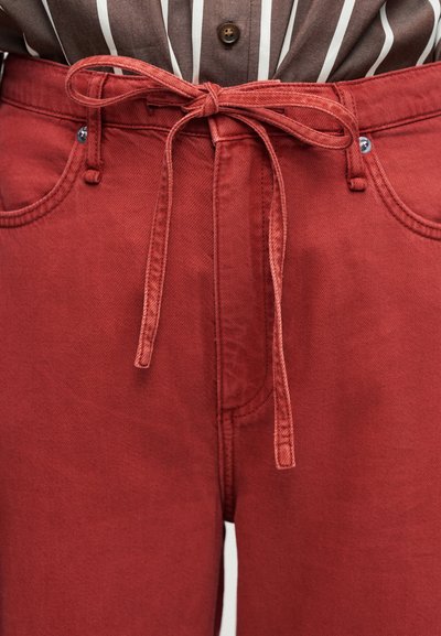 Κοντινό πλάνο κόκκινων παντελονιών με ψηλή μέση, υφασμάτινη ζώνη δεμένη, ασημένιες τρουκ και καφέ ριγέ πουκάμισο που έχει μπεί μέσα.