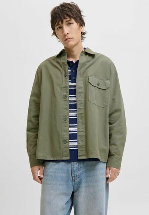 Young person with short brown hair wearing an olive green button-up shirt over a navy striped sweater and light blue jeans.