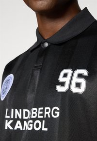 Sort poloshirt med en subtil glans, har et hvidt nummer "96", mærket "LIND/BERG KANGOL" og et cirkulært logo-patch.