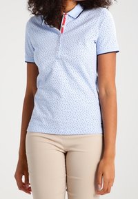 Ljusblå pikétröja med små röda och marinblåa prickar, krage och korta ärmar. Har en knappslå och marinblåa detaljer.