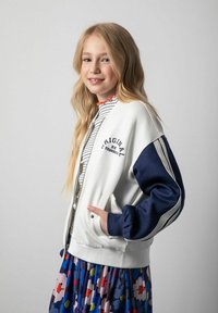 Biały bomber z granatowymi rękawami z napisem "Bądź oryginalny". W połączeniu z paskowanym topem i spódnicą w kwiatowy wzór.