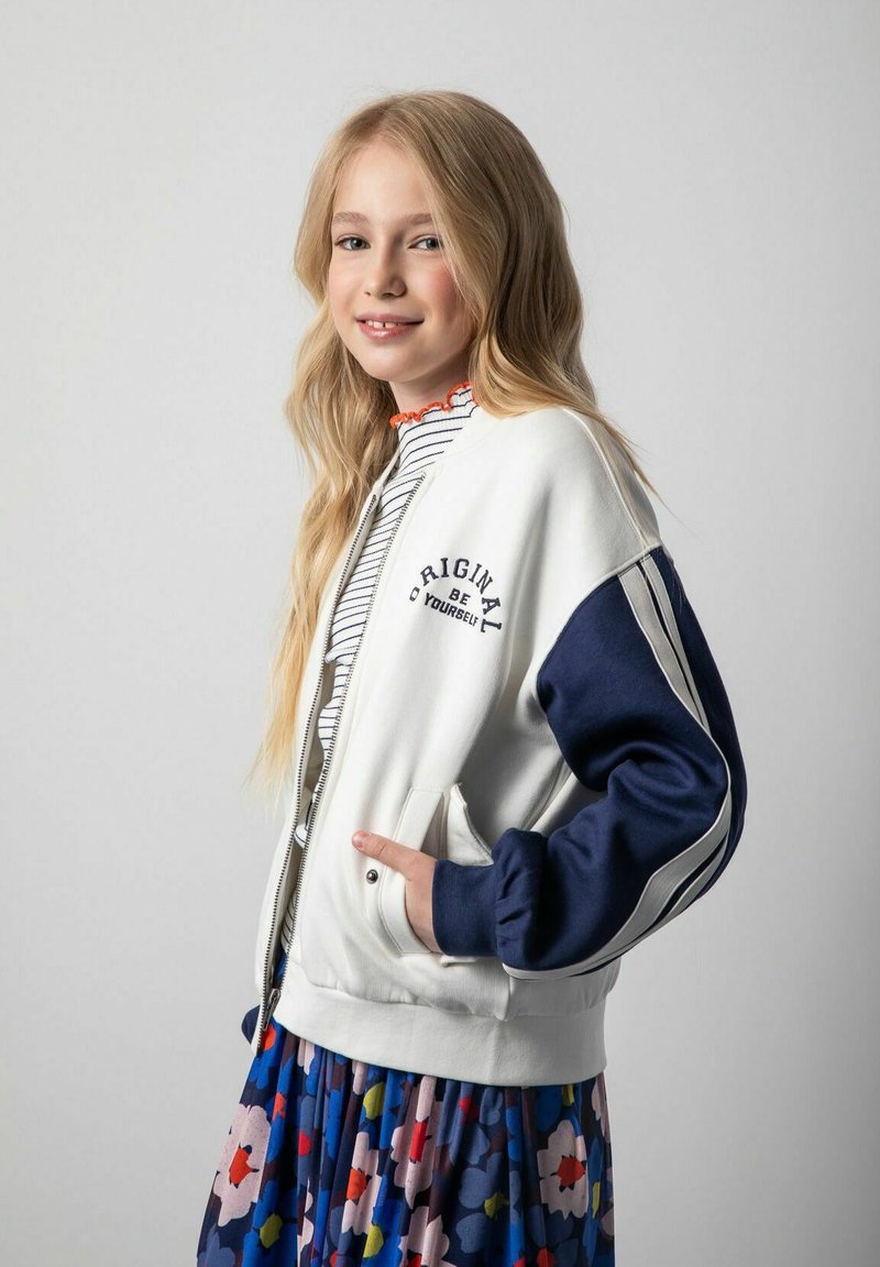 Biały bomber z granatowymi rękawami z napisem "Bądź oryginalny". W połączeniu z paskowanym topem i spódnicą w kwiatowy wzór.