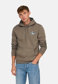 Sudadera marrón con bolsillo canguro, que presenta un gráfico de escorpión en azul y blanco en el pecho, combinada con jeans de color azul claro.