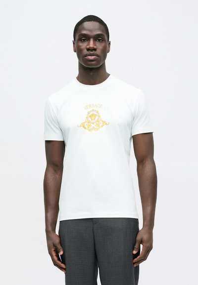 Hvid t-shirt med et gyldent Versace-logo grafisk placeret i midten. Korte ærmer og rund halsudskæring. Glat tekstur.