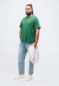 Άνδρας με dreadlocks που φοράει πράσινο μπλουζάκι, ανοιχτόχρωμο μπλε τζιν, λευκά αθλητικά παπούτσια, κρατώντας ένα διπλωμένο ριγέ πουκάμισο σε απλό φόντο.