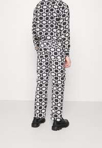 Person som bär en svart och vit retro geometrisk mönstrad långärmad skjorta och matchande byxor med svarta kraftiga skor, stående mot en vit vägg.