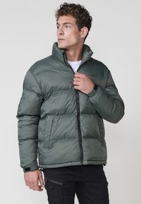Chaqueta acolchada en verde oscuro con un alto cuello, cremallera frontal y dos bolsillos laterales. Textura suave con un patrón acolchado y detalles en negro.