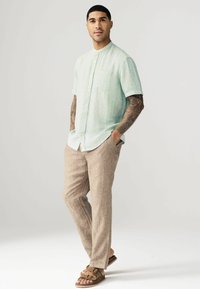 Camisa de botón de manga corta verde claro con un bolsillo en el pecho, combinada con pantalones de lino beige y sandalias marrones de slid.
