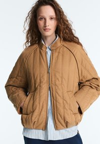 Femme aux cheveux bouclés portant une veste matelassée couleur beige à fermeture éclair par-dessus une chemise rayée, mains dans les poches de la veste, sur un fond blanc uni.