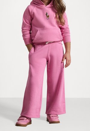 Bambino che indossa una felpa con cappuccio rosa con un logo di un cavallo, pantaloni a gamba larga abbinati e scarpe da ginnastica rosa, in piedi contro uno sfondo semplice.