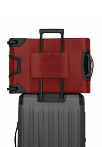 Maleta de tela roja con detalles negros y un asa retractable, situada sobre una maleta rígida de color gris texturizado.