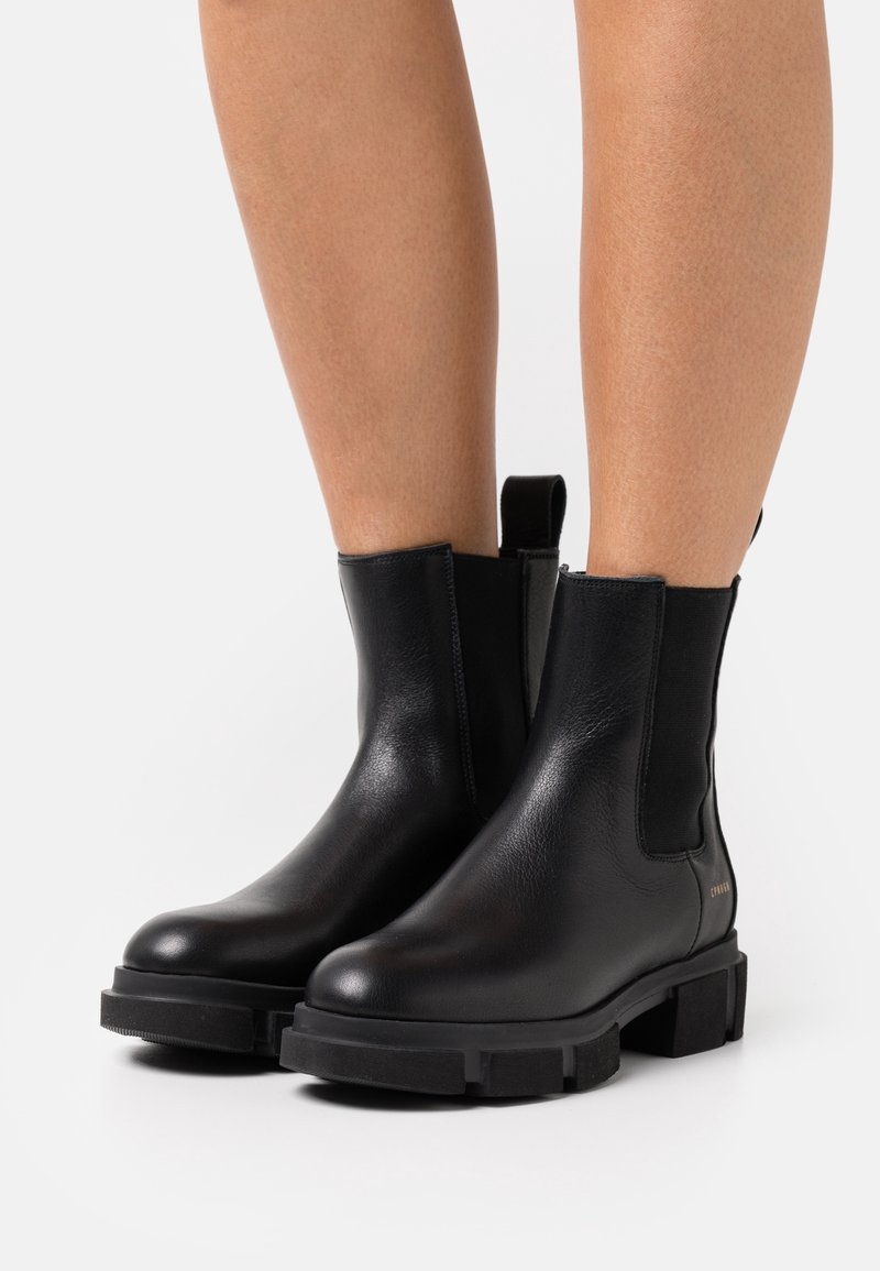 Bottes cheville en cuir noir à semelles épaisses portées sur des jambes nues sur un fond blanc uni.
