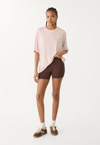 Světle růžové oversized tričko, hnědé přiléhavé šortky, hnědé tenisky a bílé kotníkové ponožky. Hladký materiál s uvolněnou siluetou, ležérní design.