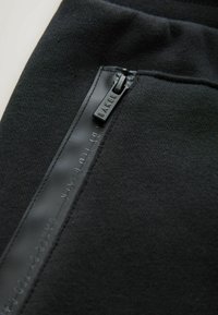 Fekete kapucnis pulóver anyaga, amely egy matt fekete cipzárhúzóval ellátott cipzáras zsebet tartalmaz, amelyen a "BAKER" felirat dombornyomott, a varrások mentén pedig sima textúrájú.