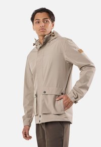 Mand iført beige hættejakke med forlommer, stående med den ene hånd i lommen, kigger let til siden.