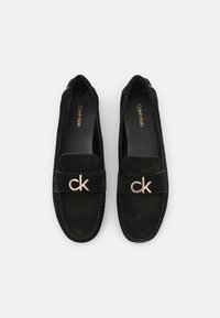 Mocassins en daim noir à semelle plate, avec une large bride et un logo « ck » doré. Texture lisse avec des détails de couture contrastants.