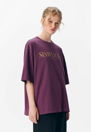 Losse fit, korte mouwen t-shirt in diep paars katoen. Bevat gouden tekst "SIMPLICITY" op de voorkant. Rechte onderkant.