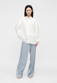 Femeie stând cu mâna pe șold, purtând o cămașă albă largă cu nasturi și pantaloni largi bleu deschis pe un fundal simplu.