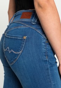 Blå denimjeans med en slät textur, hög midja, subtila sömnadsdetaljer och en brun läderbakpatch med logotyp. Bakfickan har ett vågmönster.