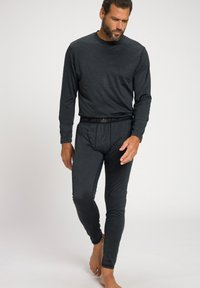 Mann mit Bart, der ein langärmliges, dunkelgraues Oberteil und passende thermische Leggings trägt, barfuß vor einem einfachen hellen Hintergrund geht.