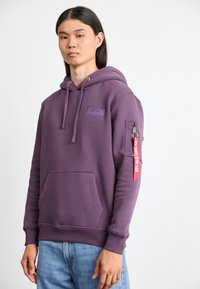 Sudadera morada de mezcla de algodón, con un bolsillo frontal, capucha con cordón y un bolsillo lateral con cremallera que tiene una etiqueta roja. Textura suave.