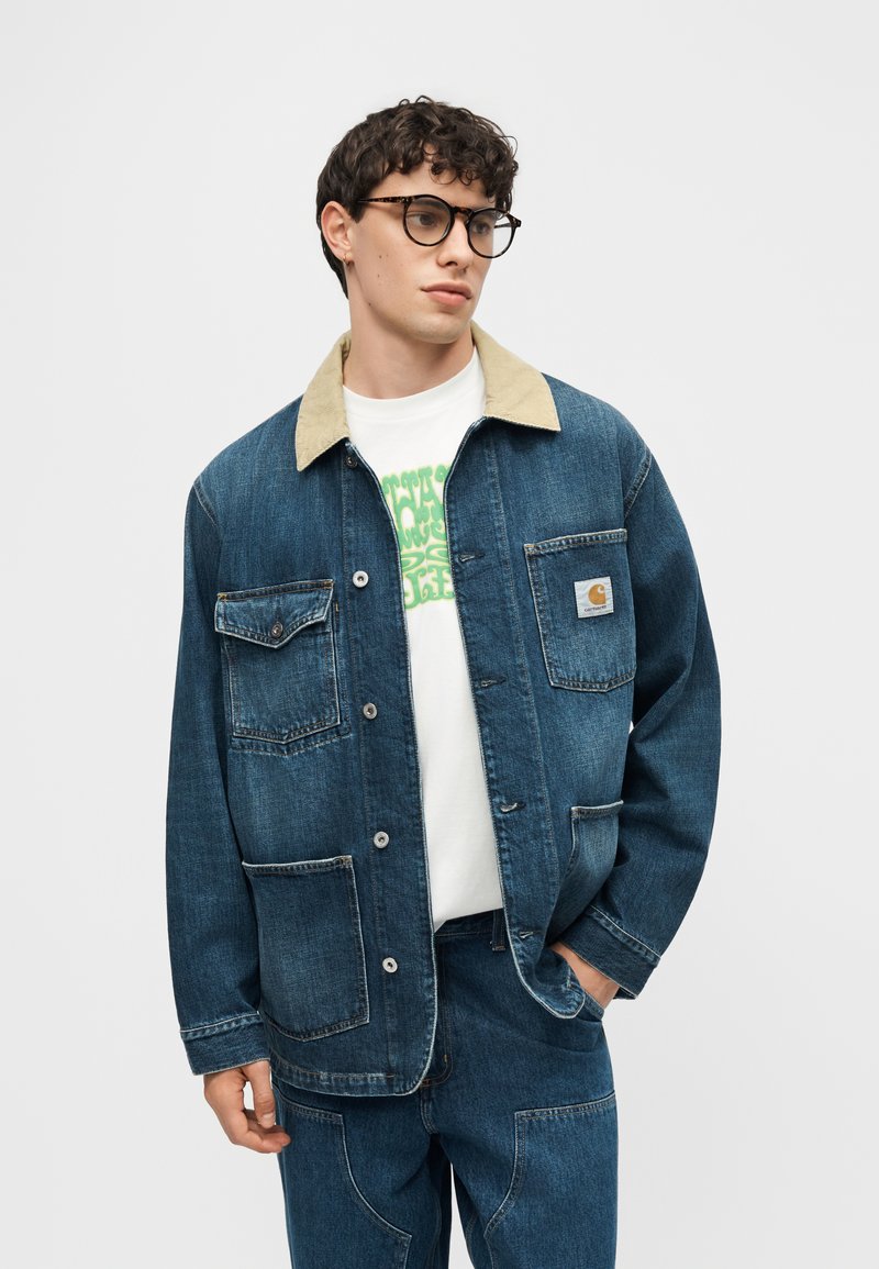 Jonge man met schildpadbril, blauwe spijkerjas met beige kraag, wit grafisch T-shirt en bijpassende blauwe spijkerbroek.