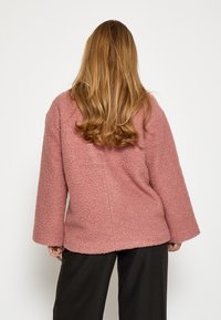 Mīksts rozā garas piedurknes džemperis ar vaļīgu piegriezumu un platām piedurknēm, ar teksturētu virsmu un noapaļotu apakšmalu, apvienots ar melnām biksēm.