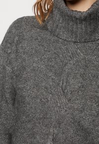 Suéter gris de punto con un alto cuello acanalado y un patrón de trenzas en la parte delantera. Tejido texturizado con un acabado suave.