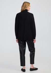 Kobieta zwrócona plecami, ubrana w czarny sweter z wysokim kołnierzem, czarne spodnie z imitacji skóry i czarne płaskie buty, na jednolitym szarym tle.