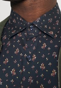 Kortärmad knäppt skjorta i mörkt tyg med ett upprepat blommönster i rött, vitt och grönt. Blåa knappar vid kragen.
