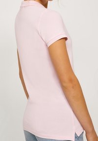 Žena v světle růžovém polo tričku s krátkým rukávem a světle modrých džínách, zobrazená zezadu a mírně otočená na stranu.