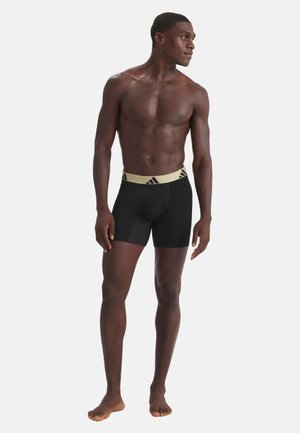 Athletischer Mann, barfuß stehend, oberkörperfrei, trägt schwarze Boxer-Shorts mit einem beigefarbenen Bund und schwarzem Logo.
