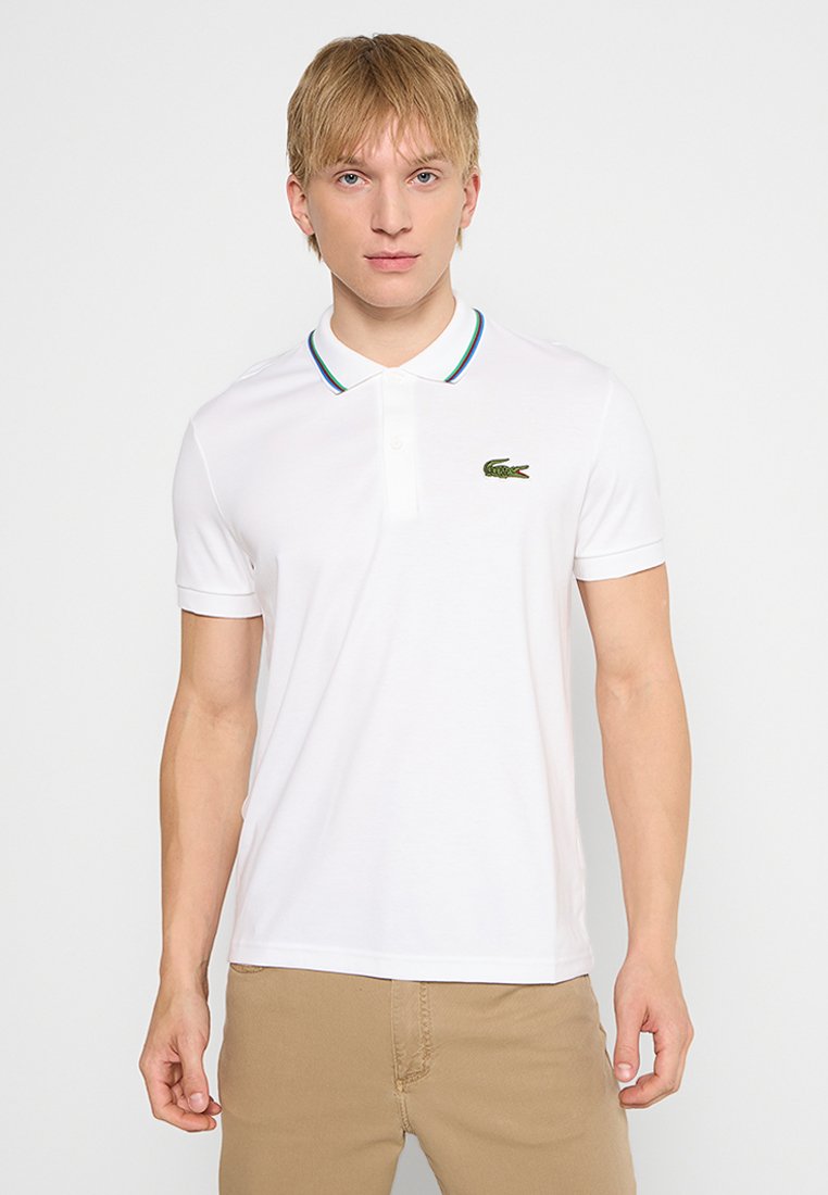 Chemise polo blanche avec un col rayé vert et bleu, à manches courtes, et un logo crocodile Lacoste vert sur le côté gauche.