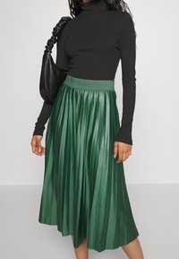 Nainen, jolla on päällään musta pitkähihainen ribbipaita, vihreä pliseerattu midi-hame ja joka kantaa mustaa rypytettyä olkahamstria.